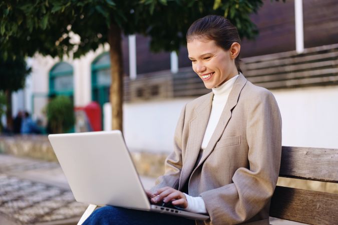 A Young Woman Happily Working on Her Laptop Outdoors While Sitting on a Bench in the Park