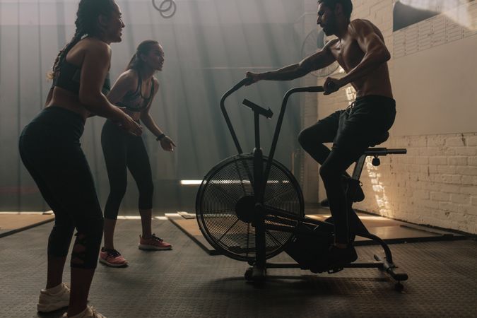 Two women motivating man exercising on air bike in gym
