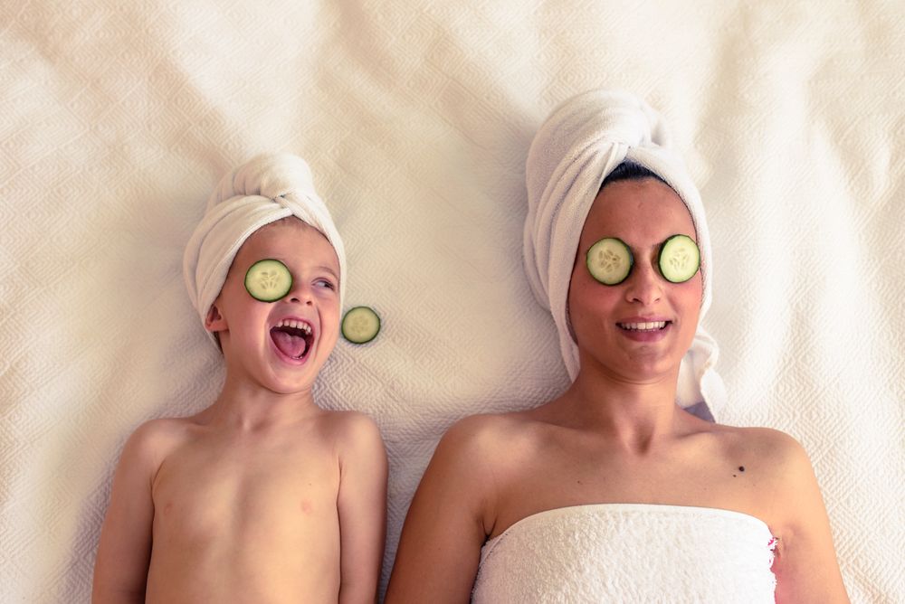 Cheerful kid with mother with cucumber on eyes
