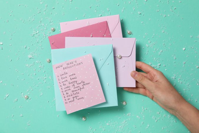 Paper with list, envelopes and female hand on green background, top view