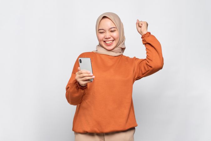 Muslim woman winning something in headscarf and orange shirt holding phone