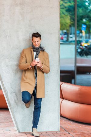 Smiling man leaning on wall outside texting on phone and wearing a camel coat 
