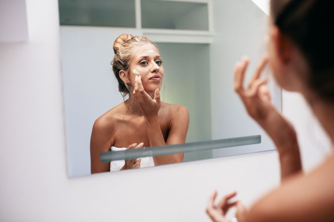 Young woman reflection in mirror applying cream in bathroom