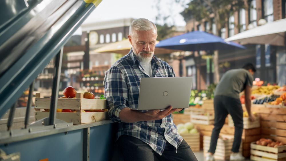 Businessman Working on Laptop Computer, Contacting Farm Suppliers About Fresh Produce for a Sunday Market Event. Male Sitting Next to a Street Vendor Booth with Organic GMO-Free Produce