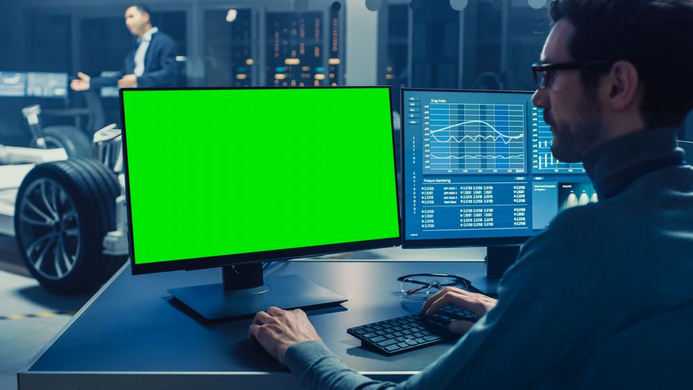 Professional Engineer Works on a Computer with Green Screen Mock Up and Tests the Electric Car Chassis Prototype with Wheels, Batteries and Engine Standing in a High Tech Development Laboratory.