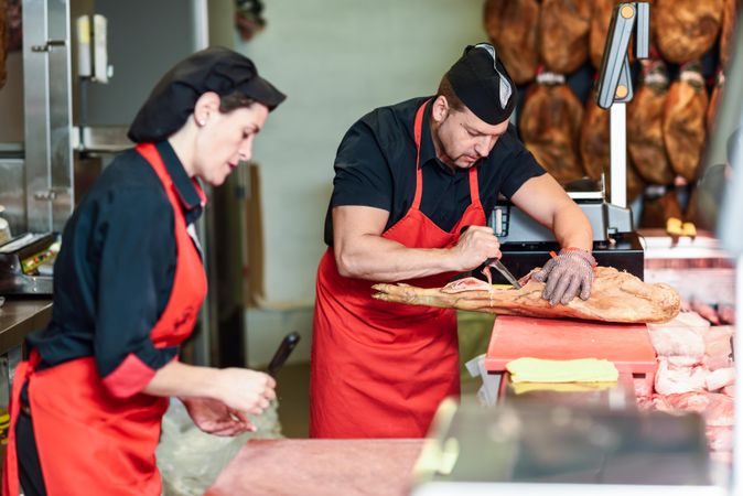 Butcher cutting cured ham  with knife with female colleague standing next to him