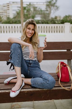 Happy woman with disposable cup of coffee sitting on street bench