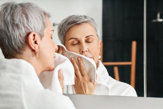 Woman with short grey hair drying her face in mirror