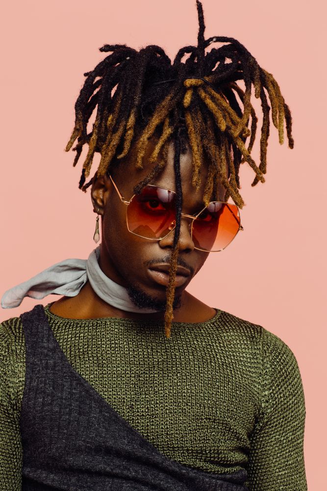 Portrait of a serious young man in green with dreadlocks and red sunglasses, isolated on pink