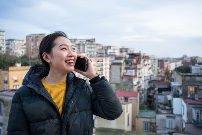 Joyful Asian female tourist in warm wear calling on smartphone at big old city