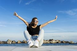 Woman with long hair sitting on promenade against blue sky while looking away with a big smile