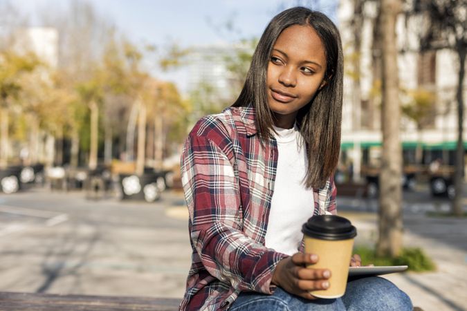 Female in plaid shirt sitting outside with tablet and to go beverage