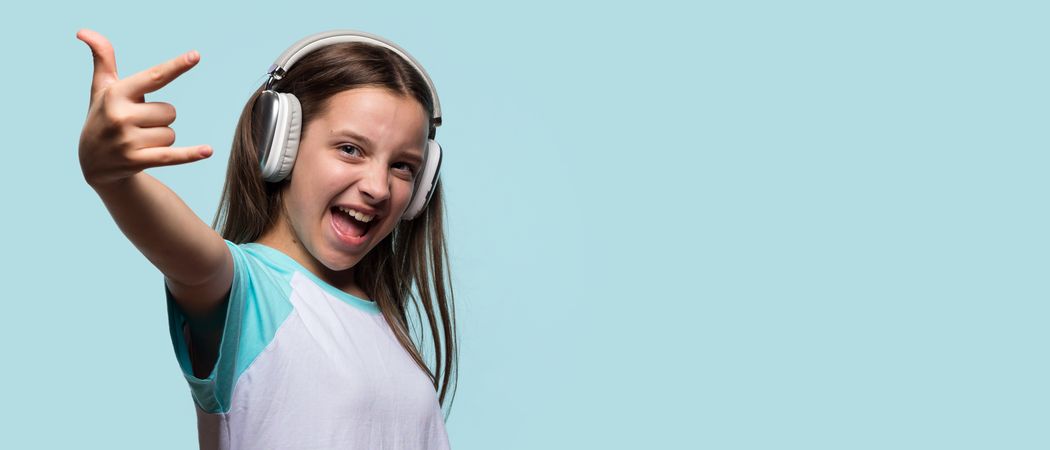 Cheerful girl wearing headphones, enthusiastically showing rock hand gesture.