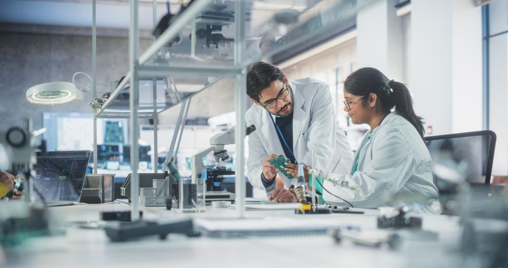 Two Diverse Multiethnic Colleagues Chatting in Laboratory, Working on a Circuit Board for a Computer.