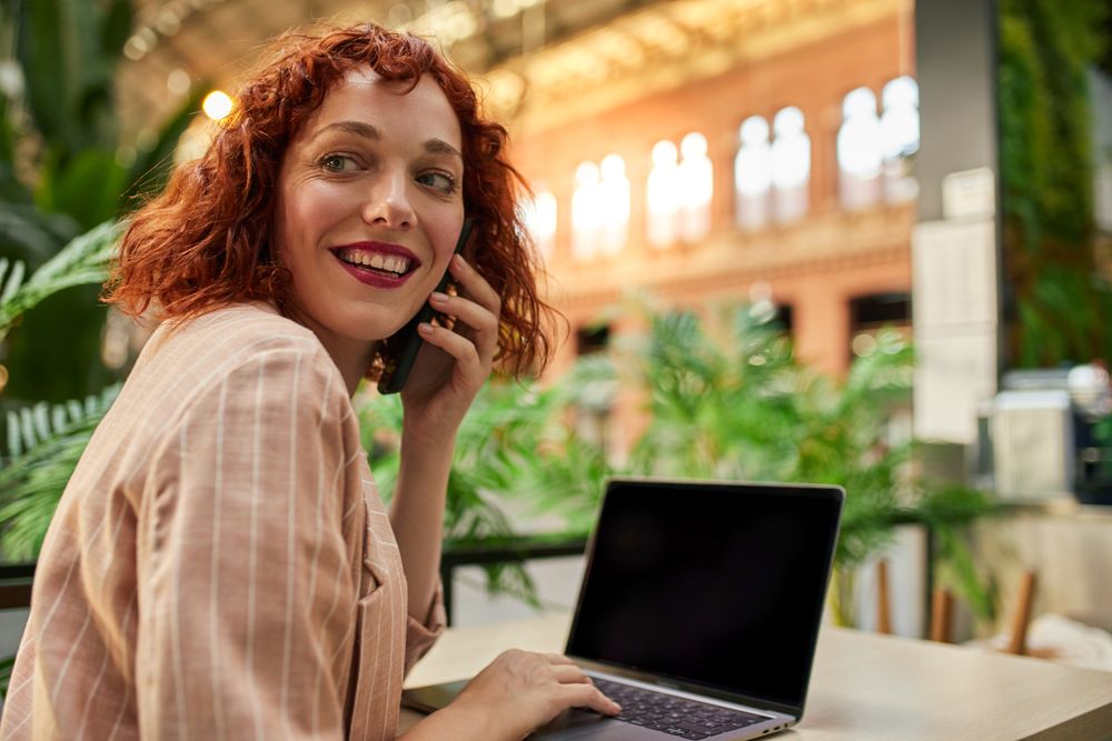 Cheerful young woman working on laptop in cafe and talking phone