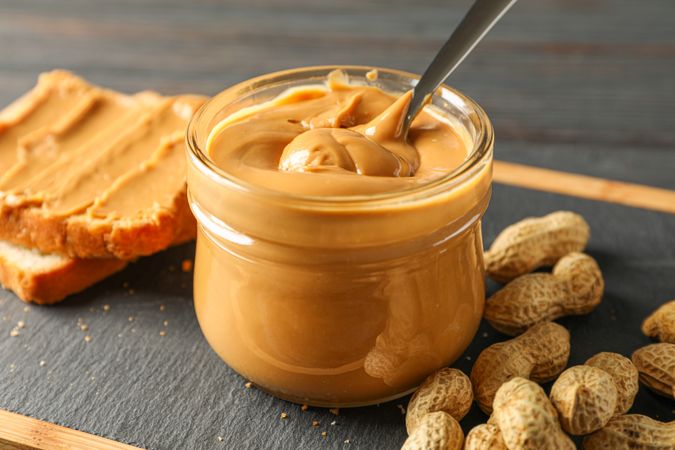 Glass jar with peanut butter and spoon, peanut, peanut butter sandwich and cutting board on wooden background, space for text and closeup