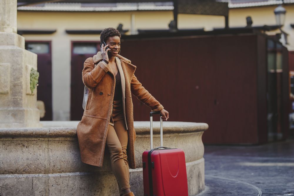 Stylish Black woman talking on smartphone with luggage on street