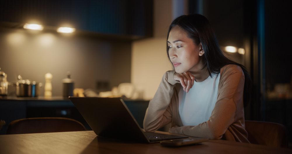 Authentic Asian Woman Sitting at a Table in Stylish Kitchen and Using Laptop Computer at Home at Night.