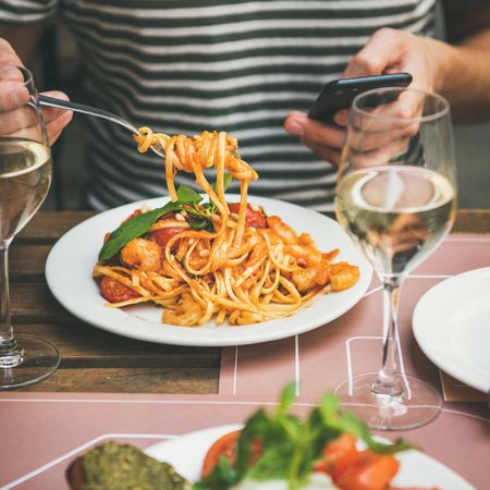 Man in striped shirt eating pasta with fork, wine, and smartphone