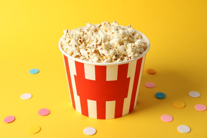 Confetti and bucket with popcorn on yellow background, copy space
