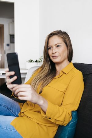 Beautiful adult female sitting on sofa while texting on smartphone