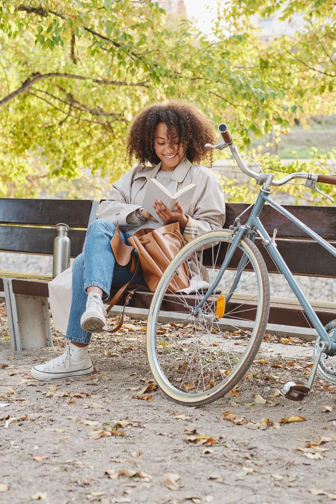 Cheerful Black woman reading book on bench near bicycle in park