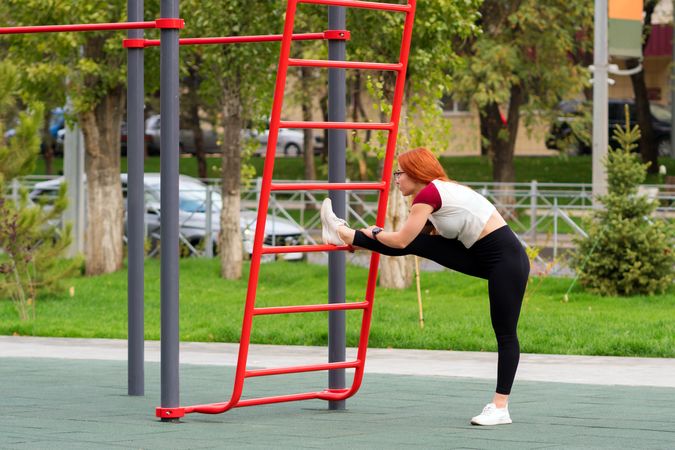 Woman stretching leg on outdoor fitness equipment in park.