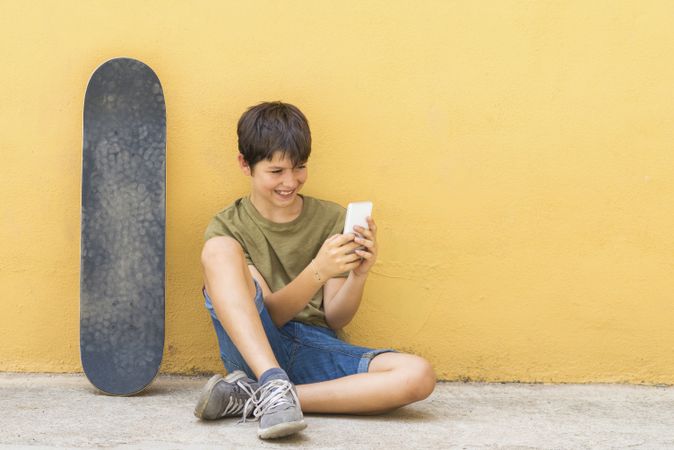 Teenage boy sitting on ground leaning on a yellow wall and texting