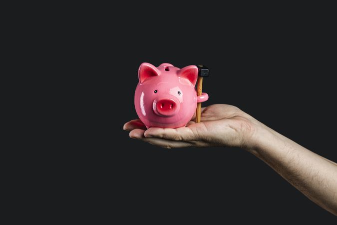 Hand holding a pink piggy bank with a hammer in it, on a dark background.