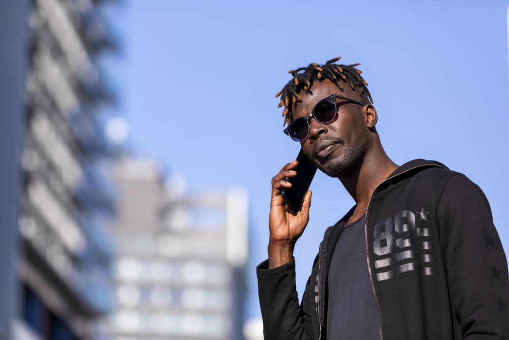Serious Black man with sunglasses standing against cityscape on the street while using a mobile phone in sunny day