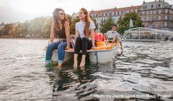 Two young female friends sitting in front of boat with their feet in the water 482Wq0