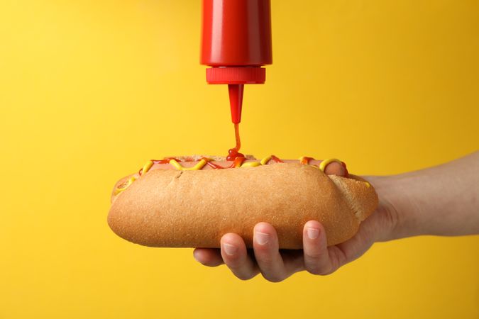 Person pouring ketchup on hot dog, on yellow background