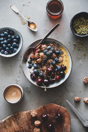 Oat granola yogurt bowl with cherries, blueberries, honey, nuts, coffee, wooden board in foreground