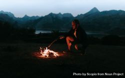 Man sitting beside bonfire in the woods near mountains and lake 423R3b