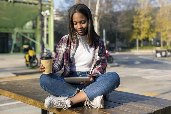 Female in plaid shirt sitting on table outside reading tablet with coffee