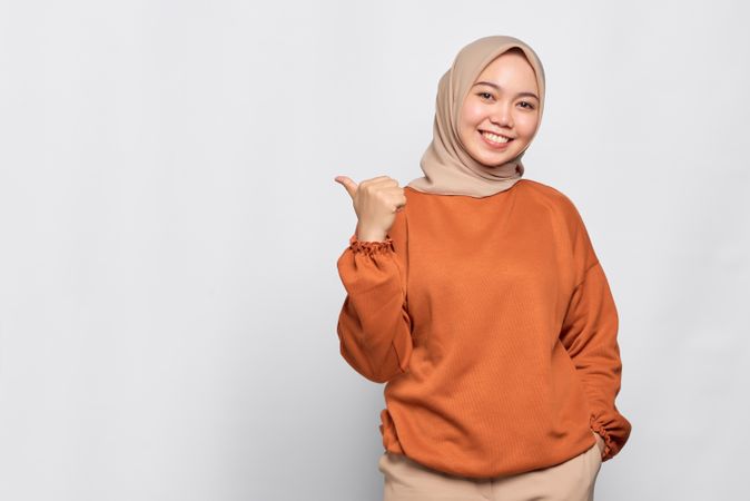 Smiling Muslim woman in headscarf and orange sweater gesturing behind her with thumb