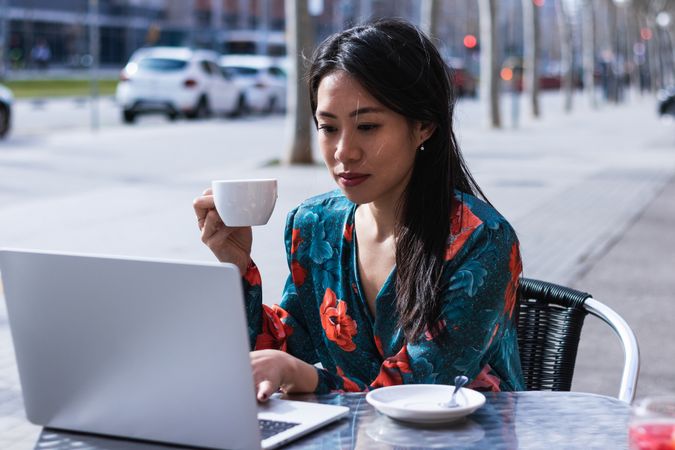Long-haired brunette Asian woman having a coffee on a terrace of a cafe while is working with a laptop