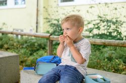 Adorable child boy sitting on bench with lunchbox and eating food in daylight