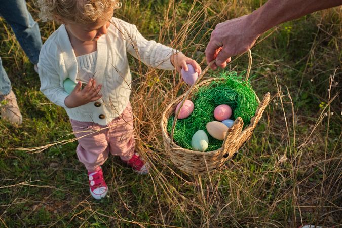 Crop girl putting Easter eggs into basket on grassy meadow