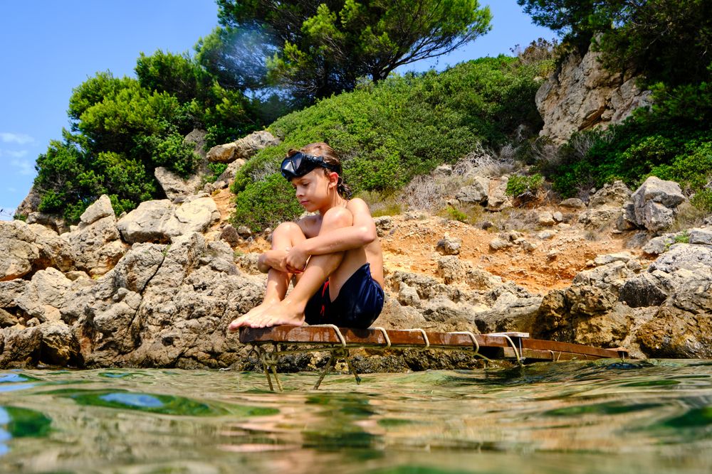 Boy in swimsuit sitting on wooden plank and looking at water