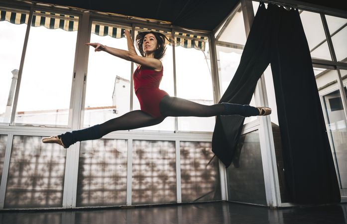 Young slim ballerina jumping above ground in studio flexing legs.
