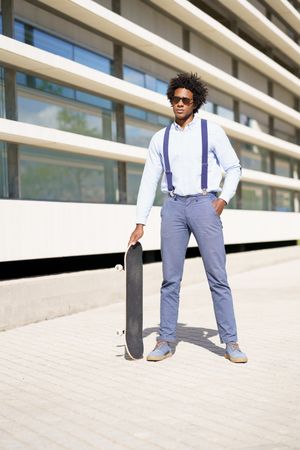 Serious man in sunglasses standing tall with skateboard outside building, vertical