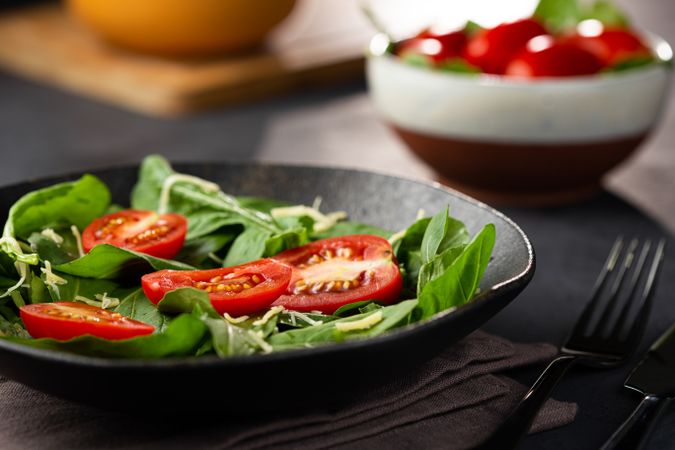 Side view of tomato and spinach salad served in dark bowl with cutlery