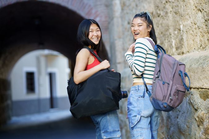 Two happy women in jeans outside looking back with bags and camera