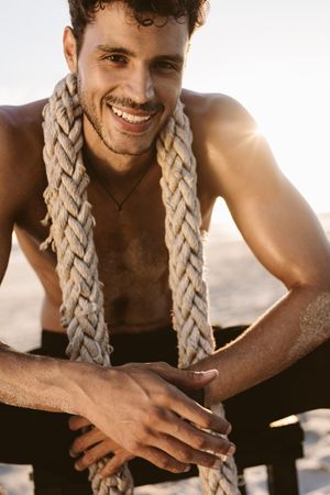 Bare chested man relaxing after workout at a beach with a battle rope around his neck