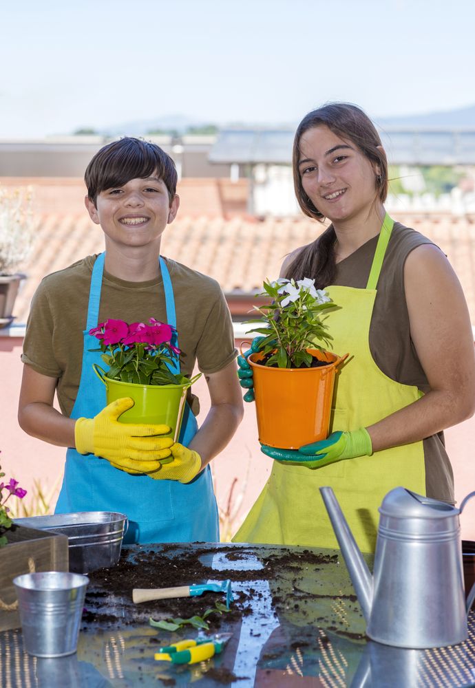 Two smiling teenager wearing gardener apron posing with plants on balcony
