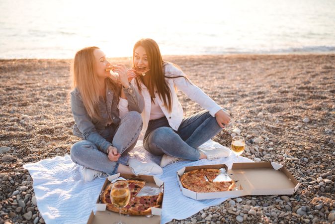 Two women eating pizza and sitting on sandy seashore