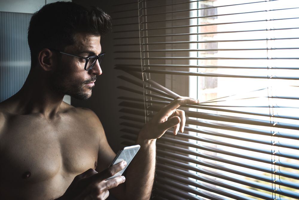 Man standing by a windows blind on the mobile phone