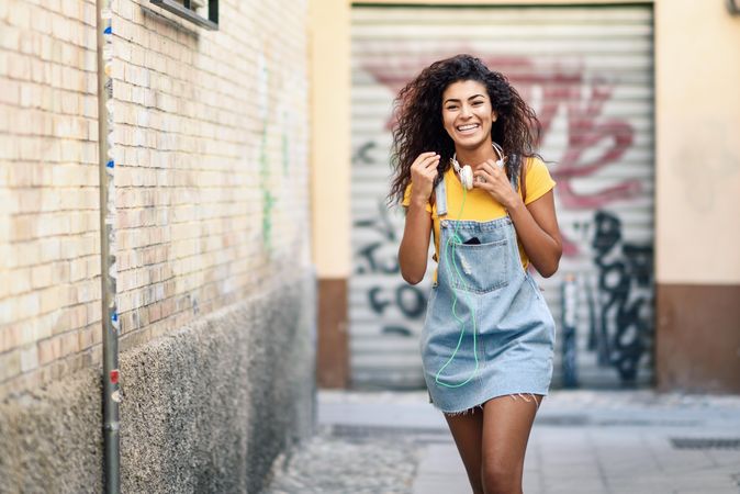 Arab woman with headphones and curly hairstyle walking outdoors in lane with graffiti