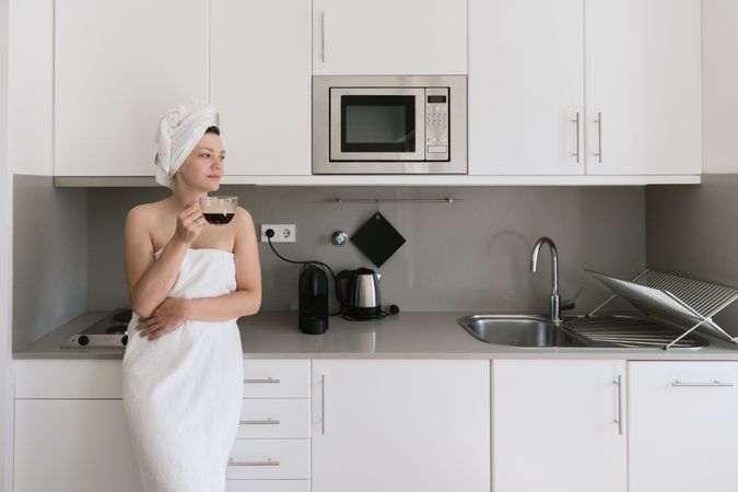 Joyful adult woman enjoying day off while having hot drink after bath in apartment with simple interior design